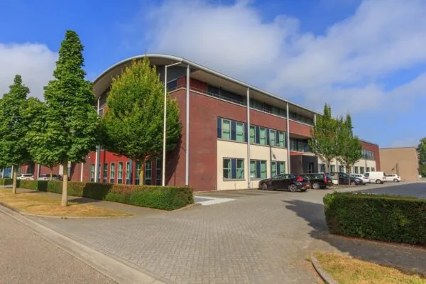 Modern kantoorgebouw De Wel met parkeerplaatsen en omliggend groen op een zonnige dag.